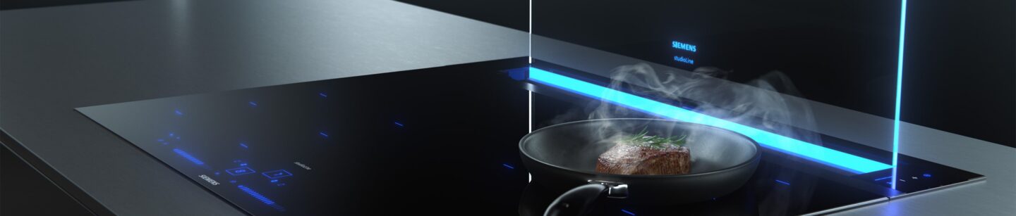 Bild zeigt einen Herd auf dem eine Bratpfanne steht. Im Hintergrund ist der integrierte Dunstabzug ausgefahren und zieht den Kochdampf ein.